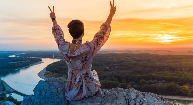 Une femme surplombant un paysage face au soleil couchant faisant le signe de victoire avec ses mains