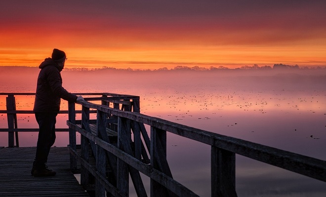 Un homme sur un ponton en bois en train de contempler un lac brumeux au lever du soleil
